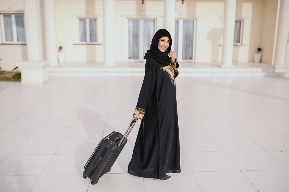 smiling-muslim-woman-traditional-wear-standing-front-her-house-holding-luggage